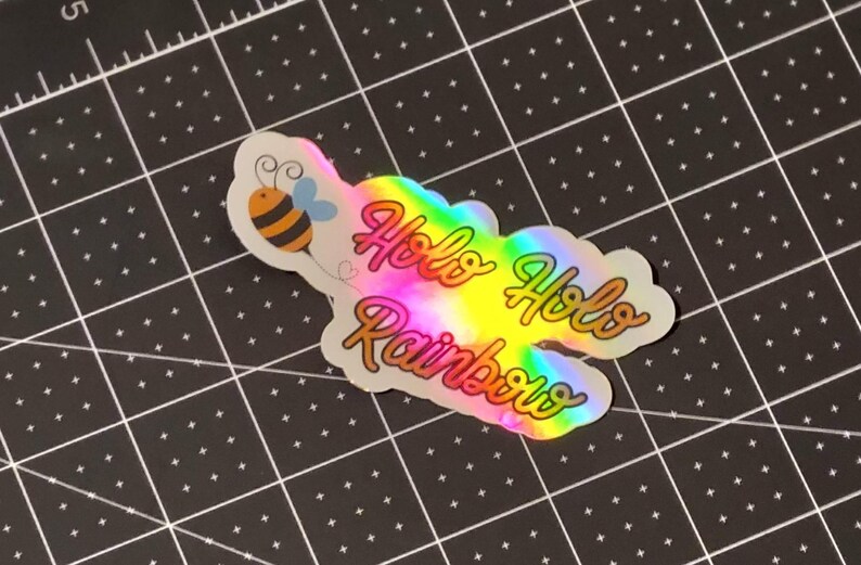 Puede incluir: Una pegatina hologr&aacute;fica con las palabras "Holo Holo Rainbows" en una fuente de escritura. La pegatina tiene un degradado de arco&iacute;ris y una ilustraci&oacute;n de abeja de dibujos animados. La pegatina est&aacute; sobre una esterilla de corte negra.