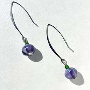 May include: A pair of drop earrings featuring silver-toned curved ear wires. Each earring displays a small, round, purple and white marbled bead accented by a tiny, vibrant green glass bead at the top, creating a simple and balanced jewelry design.