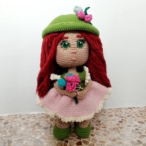 May include: A handmade crochet doll with red hair, green eyes, and a green hat adorned with pink flowers. The doll wears a pink dress with a white trim and holds a bouquet of colorful crochet flowers. The doll's shoes are green.