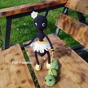 May include: A black crochet doll with a white daisy-like skirt and a matching flower on its head. It holds a green caterpillar toy on a yellow string. The doll has yellow shoes and is sitting on a wooden bench.