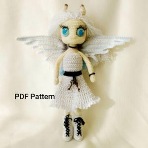 Puede incluir: Muñeca de ganchillo hecha a mano con diseño de hada. La muñeca tiene cabello blanco, ojos azules y alas delicadas. Viste un vestido blanco con cinturón negro y dijes de llaves. El texto "PDF Pattern" es visible.
