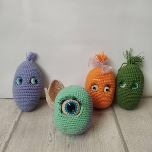 May include: A collection of five handmade crochet creatures in various colors. The creatures have embroidered eyes and some have decorative elements like a tassel or ribbon. The color palette includes purple, orange, green, and teal.