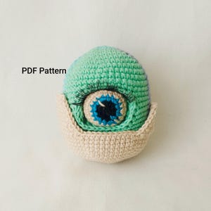 May include: A crocheted eye-shaped plush toy in shades of teal, blue, and beige. The eye features a blue iris with a black pupil, surrounded by a teal and beige border. Long, black eyelashes are attached above the eye. The text "PDF Pattern" is visible.