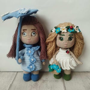 Op de afbeelding: Twee handgemaakte gehaakte poppen. De ene pop draagt een lichtblauwe jurk, laarzen en houdt een blauwe paraplu vast. De andere pop heeft een witte jurk, een bloemenkrans en draagt een mandje met blauwe bloemen. Beide poppen hebben lang haar.