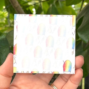May include: Square notepad with a repeating pattern of rainbow-colored shaved ice treats. The notepad is held in a hand, with a gold ring visible. The background is blurred green foliage.