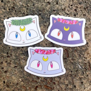 Può includere: Tre adesivi con disegni di gatti dei cartoni animati con pelliccia di diversi colori e corone di fiori. I gatti hanno lune crescenti gialle sulla fronte e occhi rosa e blu. Gli adesivi sono su uno sfondo grigio testurizzato.