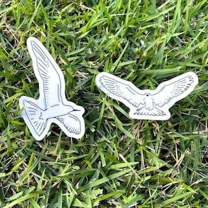 May include: Two white stickers with black line drawings of birds in flight. The birds are facing different directions.