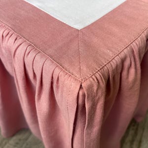 May include: Close-up of a dusty rose bed skirt with a pleated design. The skirt has a tailored corner and a white top border. The fabric appears to be linen or a similar woven material, with visible texture.