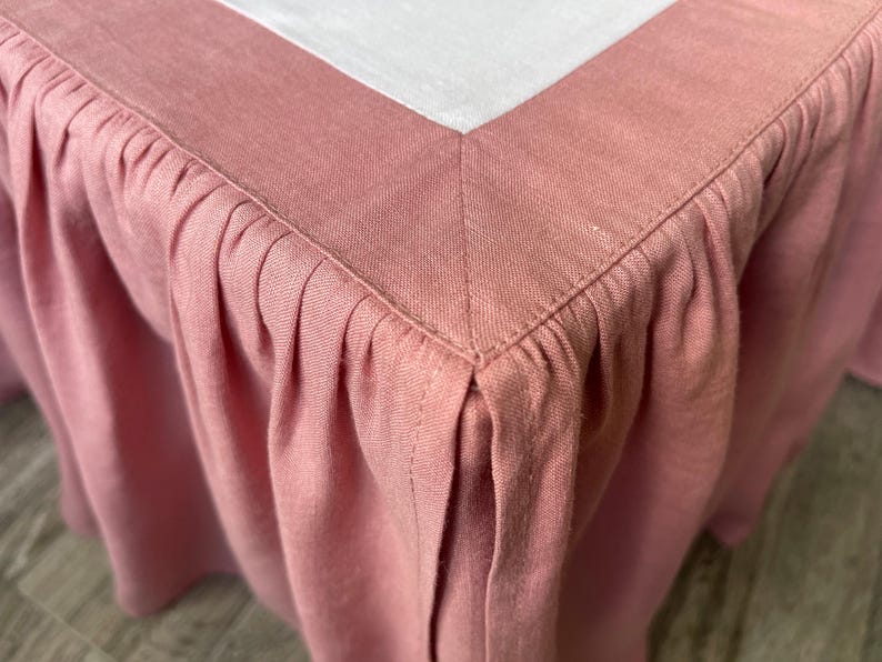 May include: Close-up of a dusty rose bed skirt with a pleated design. The skirt has a tailored corner and a white top border. The fabric appears to be linen or a similar woven material, with visible texture.