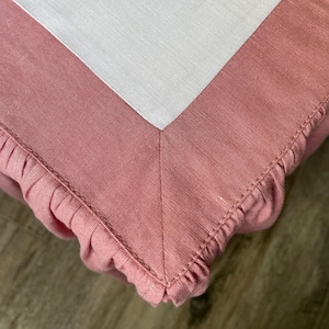 May include: Close-up of a pink linen fabric with a white linen corner. The pink fabric has a ruffled edge and is stitched together. The fabric appears to be a tablecloth or a similar textile.
