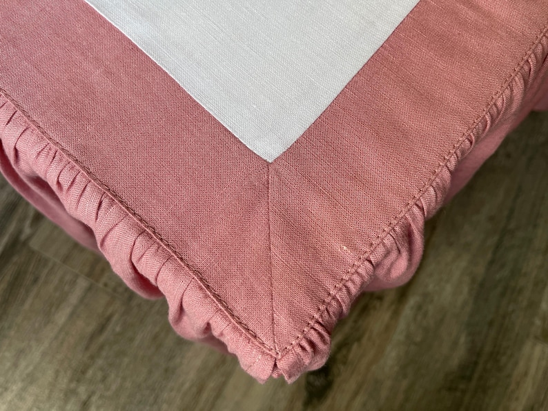 May include: Close-up of a pink linen fabric with a white linen corner. The pink fabric has a ruffled edge and is stitched together. The fabric appears to be a tablecloth or a similar textile.