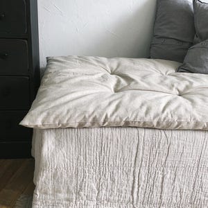 Può includere: Un cuscino da pavimento rettangolare color beige con una morbida fodera in tessuto trapuntato. Il cuscino presenta una parte superiore spessa e imbottita con bottoni a vista e una base strutturata con cuciture che arriva fino al pavimento.