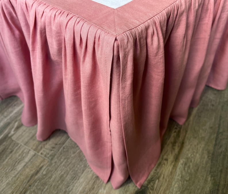 May include: Close-up of a dusty rose bed skirt with a pleated design. The fabric is a solid color and appears to be linen or a similar material. The skirt is tailored with a corner split.