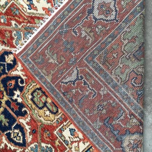May include: Close-up of a section of a traditional rug with intricate patterns. The rug features a rich color palette of red, navy blue, cream, and green. The design includes geometric shapes and floral motifs, with a detailed border and fringe.