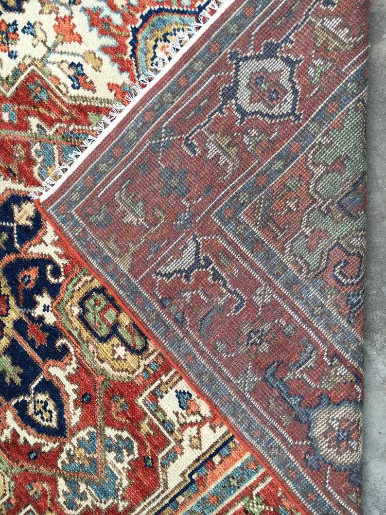 May include: Close-up of a section of a traditional rug with intricate patterns. The rug features a rich color palette of red, navy blue, cream, and green. The design includes geometric shapes and floral motifs, with a detailed border and fringe.
