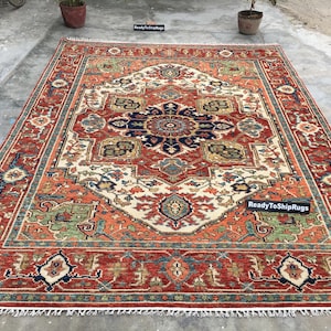 May include: A large, rectangular rug with an intricate, symmetrical design. The rug features a central medallion in navy blue and cream, surrounded by a red border with green and blue accents. The text "ReadyToShipRugs" is visible.