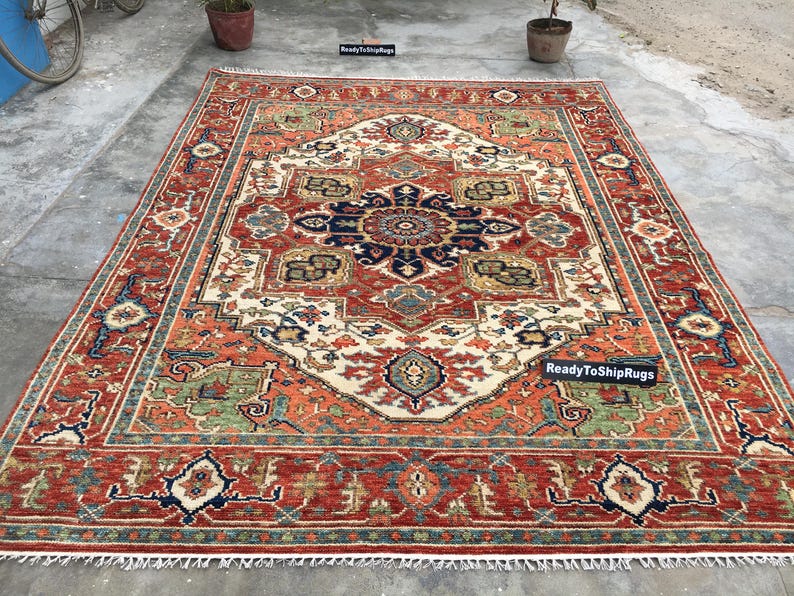 May include: A large, rectangular rug with an intricate, symmetrical design. The rug features a central medallion in navy blue and cream, surrounded by a red border with green and blue accents. The text "ReadyToShipRugs" is visible.
