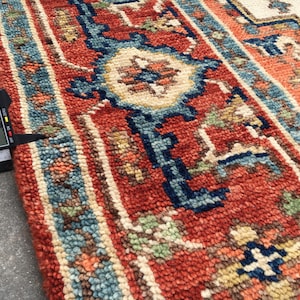 May include: Close-up of a hand-knotted rug with a traditional design. The rug features a red background with blue, cream, and green geometric patterns. The rug's thickness is measured at 0.7 inches.