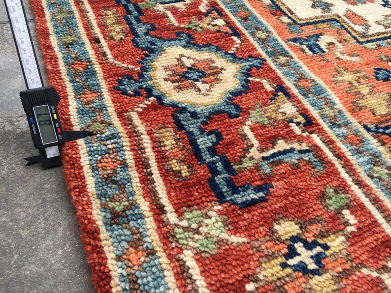 May include: Close-up of a hand-knotted rug with a traditional design. The rug features a red background with blue, cream, and green geometric patterns. The rug's thickness is measured at 0.7 inches.