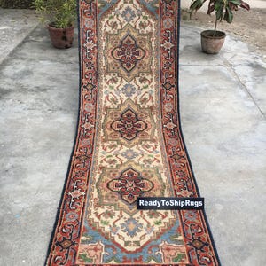 May include: A rectangular rug with an intricate, symmetrical design. The rug features a central cream-colored field with red, blue, and brown geometric patterns. The border is a deep navy blue with a repeating floral motif. The rug has a fringe on both ends.