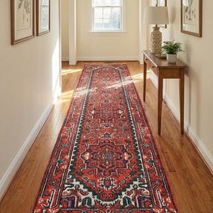 May include: A long, rectangular red rug with an intricate geometric pattern, featuring navy, green, and white accents. The rug is placed in a hallway with hardwood floors and framed artwork on the walls. The rug has a fringed edge.
