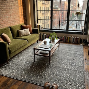 May include: A rectangular area rug featuring a dark gray base with a repeating pattern of small, light-colored dots. The rug has a textured, woven appearance and is positioned on a wooden floor, partially covered by a wooden coffee table and a sofa.