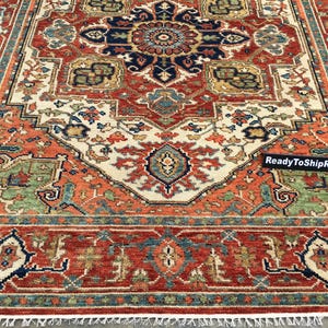 May include: A rectangular rug with a detailed, symmetrical design. The rug features a central medallion in navy blue and rust-red, surrounded by cream, green, and gold patterns. The border is a deep red with intricate motifs, and the text "ReadyToShipRugs" is visible.