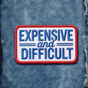 May include: A rectangular patch with the words "EXPENSIVE and DIFFICULT" in blue, embroidered on a white background. The patch has a red border and is set against a blue denim background.