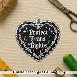 May include: Black heart-shaped patch with white text reading "Protect Trans Rights." The patch is bordered by barbed wire and a sword. Additional elements include a pair of scissors, thread, and a measuring tape.