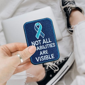 May include: A navy blue embroidered patch with a teal ribbon and the text "NOT ALL ABILITIES ARE VISIBLE". The patch is rectangular with rounded corners. A silver ring is visible on a finger.