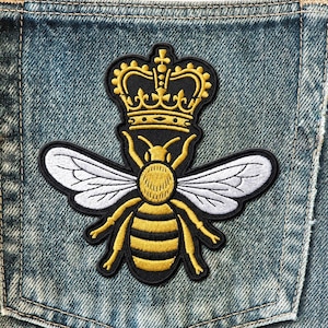 May include: Embroidered patch featuring a bee wearing a gold crown. The bee has white wings, a yellow body with black stripes, and yellow legs. The patch is on a denim background.