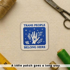 May include: Square patch with a blue background and white text that reads "TRANS PEOPLE BELONG HERE." The patch features a white hand with a heart in the center, surrounded by stars and leaves. The patch is on a beige fabric surface.