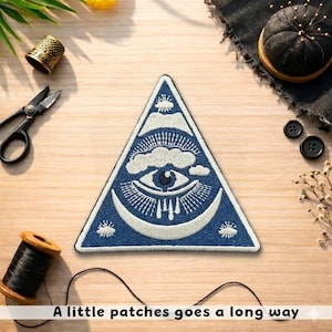May include: A blue and white embroidered patch in a triangular shape, featuring an eye, clouds, and a crescent moon design. The text "A little patches goes a long way" is at the bottom. Other sewing supplies are in the background.