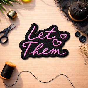 May include: A black patch with the words "Let Them" in pink cursive script, with two pink hearts. The patch is surrounded by sewing supplies, including scissors, thread, and buttons, on a light wood surface.