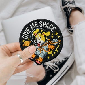 May include: A round, black embroidered patch with the text "GIVE ME SPACE" encircling a cartoon dog in an astronaut suit. The dog is yellow with orange accents, surrounded by stars and planets. The patch is held by a hand with a silver ring.