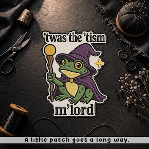 May include: Embroidered patch featuring a green frog wearing a purple wizard hat and cape, holding a staff. Text on the patch reads "'twas the 'tism m'lord" and "A little patch goes a long way."