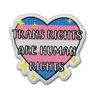 May include: A heart-shaped patch with the text "TRANS RIGHTS ARE HUMAN RIGHTS". The patch features the colors of the transgender flag: pink, white, and blue, with yellow star accents and a white border.