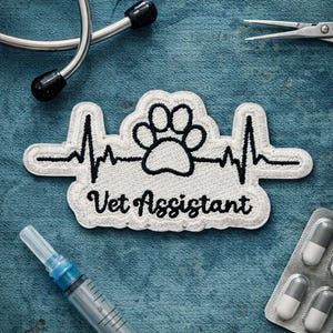 May include: A white embroidered patch with a black paw print, heartbeat line, and the words "Vet Assistant." Medical tools, including a stethoscope, syringe, and pills, are arranged around the patch on a blue denim surface.