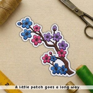 May include: Embroidered patch featuring a branch with blue, pink, and purple flowers. The patch has a white border and a brown stem. The text "A little patch goes a long way" is at the bottom.
