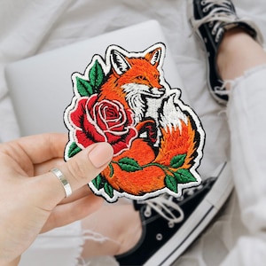 May include: Embroidered patch featuring an orange fox with a white chest and tail, nestled beside a red rose with green leaves. The patch has a white border. The patch is being held by a person.