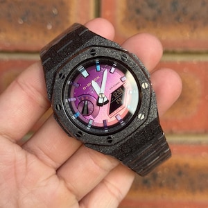 May include: A G-Shock watch with a black, glittery, textured octagonal bezel and matching link band. The watch face features a gradient purple and pink dial with silver hands, white hour markers, and a digital display showing 12:08 and WR 20BAR.