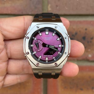 May include: A G-Shock Casio wristwatch featuring a metallic silver octagonal bezel with exposed screws, a vibrant purple dial, and a black resin strap. The watch face displays both analogue hands and a digital window showing the number 49.