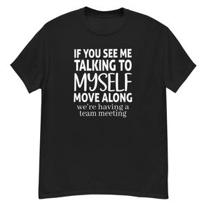 May include: Black t-shirt with white text: "IF YOU SEE ME TALKING TO MYSELF MOVE ALONG we're having a team meeting." The shirt is a short-sleeved crew neck style.