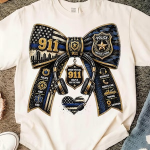 Peut inclure: Un t-shirt crème avec un grand motif de nœud au design de ligne bleue. Le nœud comprend un insigne de police, un écusson 911, un casque audio et des icônes pour la police, les pompiers, les secours, la répartition et le remorquage. Texte 911 inclus.