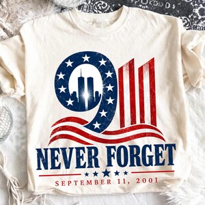 Könnte beinhalten: Ein cremefarbenes T-Shirt mit einer Grafik, die die Zahl 911, die Silhouette der Zwillingstürme und Motive der amerikanischen Flagge zeigt. Unter der Grafik steht der Text NEVER FORGET und SEPTEMBER 11, 2001 in blauen und roten Buchstaben.