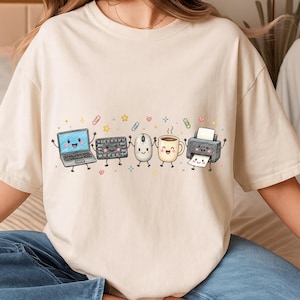 May include: A cream-colored t-shirt featuring a graphic illustration of five anthropomorphic office items holding hands: a laptop, a keyboard, a computer mouse, a coffee mug, and a printer, surrounded by small colorful paperclips, stars, and hearts.