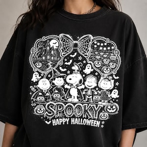 Può includere: Una t-shirt nera con una stampa grafica bianca dei personaggi Peanuts in costume di Halloween. Il design include una casa stregata, un campo di zucche, un grande fiocco, fantasmi e il testo SPOOKY HAPPY HALLOWEEN con dettagli a ragnatela.