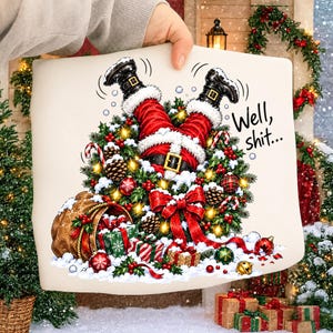 May include: A white fabric surface featuring a graphic of Santa Claus stuck upside down in a Christmas wreath. The design includes red pants, black boots, a large red bow, pinecones, ornaments, and a gift sack. The text reads, Well, shit...
