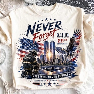 May include: A cream-colored t-shirt featuring a graphic of a kneeling firefighter, the Twin Towers, an American flag, and a bald eagle. Text includes NEVER FORGET, 9.11.01, 25TH ANNIVERSARY, WE WILL NEVER FORGET, REMEMBER, HONOR, and UNITE in red and blue.