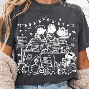 Op de afbeelding: Een antracietgrijs t-shirt met een witte afbeelding van Peanuts-personages in een busje versierd met pompoenen en lichtsnoeren. De tekst luidt PUMPKINS, COZY VIBES, SPOOKY FUN en HAPPY FALL Y'ALL. Snoopy zit op een stoel in de buurt.